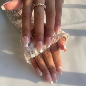 Puede incluir: Primer plano de una mano con uñas con manicura francesa. Las uñas son de un rosa natural con puntas blancas. Un anillo dorado con pequeñas piedras transparentes está en el dedo anular. La mano descansa sobre una tela de encaje blanca.