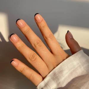 Könnte beinhalten: Eine Hand mit French-Maniküre. Die Nägel sind mit einer transparenten Basis und einer schwarzen Spitze lackiert. Die Hand liegt auf einem weißen Hintergrund.