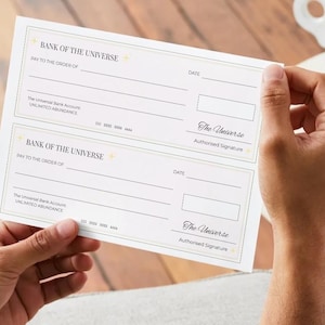May include: Two white "Bank of the Universe" checks are held by a person. The checks have lines for "Pay to the order of" and "Date". The text "The Universe" and "Unlimited Abundance" are also printed on the checks.