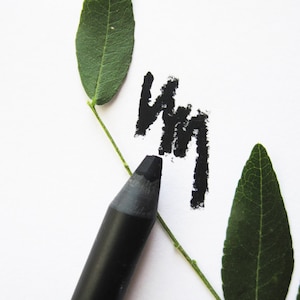 Puede incluir: Lápiz delineador de ojos negro con una rama de hoja verde en el fondo. El lápiz está sobre una superficie blanca y se ha utilizado para dibujar una línea ondulada.