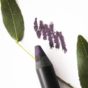 Op de afbeelding: Een zwarte eyelinerpotlood met een paarse punt, een swatch van de paarse eyeliner op een witte achtergrond en twee groene bladeren.