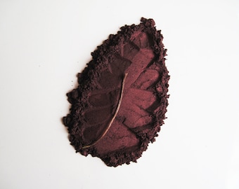 May include: A close-up of a dark red powder in the shape of a leaf. The powder is spread out on a white surface.