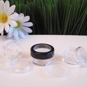 May include: Four clear plastic containers with black lids. The containers are stacked and arranged on a white surface. The containers are small and round, and they have a screw-on lid.