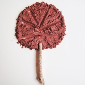 May include: A stylized image of a leaf created with a reddish-brown powder. The leaf shape has detailed vein lines and is attached to a small, light-brown twig, set against a white background. The powder has a textured appearance.