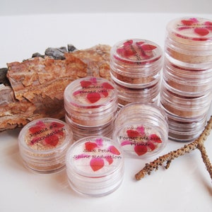 Peut inclure: Six petits pots en verre avec couvercles contenant des produits de maquillage. Les pots sont étiquetés avec les noms des produits : Pebble #2 Foundation, Flawless Tinted Veil, Forget Me Not Primer, Silk Petal Shine Reduction. Les pots sont disposés sur une surface blanche avec un morceau d'écorce en arrière-plan.