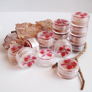 May include: A collection of small, clear jars filled with various shades of foundation and primer. The jars have labels with floral designs and text that reads "Vanilla Bark Foundation", "Forget Me Not Primer", and "Satin Foundation #4".