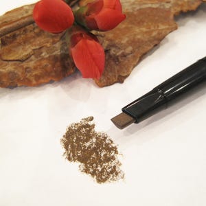 Può includere: Una matita per sopracciglia marrone con un astuccio nero. La matita è su una superficie bianca con un campione di polvere marrone accanto.