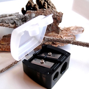 May include: A black double-hole pencil sharpener with a clear plastic lid. The sharpener has two metal blades and is designed to sharpen pencils and cosmetic pencils.