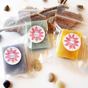 May include: Three small bars of soap in clear plastic bags. The soaps are lavender shea, mango peach, and  peppermint. Each bar has a round label with a pink flower and the name of the soap.