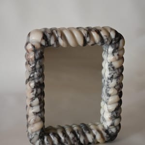 May include: A square marble mirror with a rope-like frame. The frame is white with gray and black veining. The mirror is a decorative home decor item.