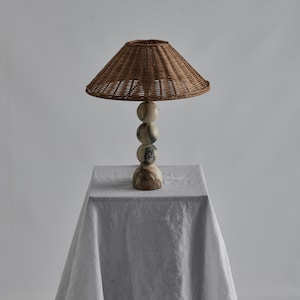 May include: A decorative table lamp with a woven rattan lampshade and a stacked marble base. The lamp has a natural color palette, with the shade and base in shades of brown and cream. The lamp is displayed on a white tablecloth.
