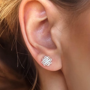 May include: A close-up of a silver stud earring with a floral design. The earring is worn on an ear, with the hair pulled back. The background is blurred with warm tones.