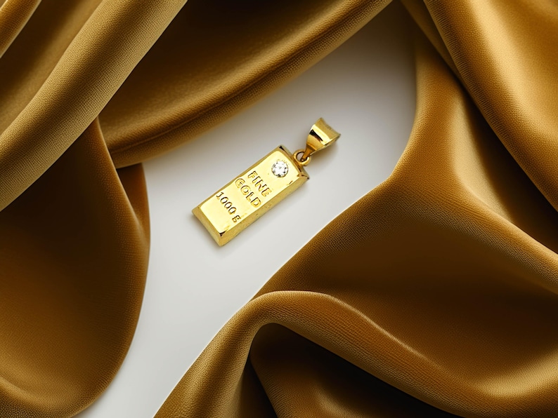May include: A gold pendant in the shape of a small gold bar, engraved with "FINE GOLD" and "1000 g". A small diamond is set into the bar. The pendant is displayed on a gold satin fabric.