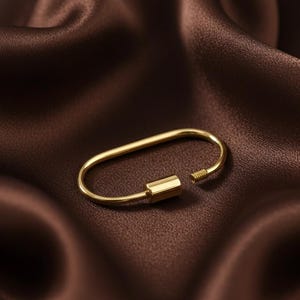 May include: A gold-coloured, oval-shaped clasp with a screw closure. The clasp is displayed on a dark brown, satin-like fabric, creating a contrast between the shiny metal and the matte background. The clasp is a jewellery component.
