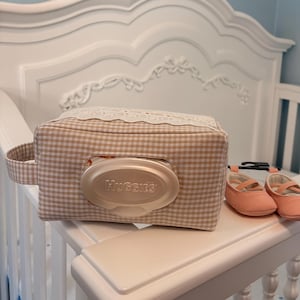 May include: A rectangular beige and white gingham fabric wipe case with a white lace trim along the top edge and a central plastic Huggies dispenser lid, accompanied by a pair of small, solid peach-colored slip-on shoes with a cross-strap design.