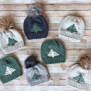 May include: A collection of knitted winter hats with a Christmas tree design. The hats are in various colors, including gray, navy, and green, and feature faux fur pom-poms. The hats are displayed on a wooden surface.