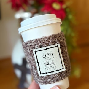 May include: A white disposable coffee cup featuring a brown crocheted sleeve. A square white patch is attached to the front of the sleeve with black stitching, displaying the text "SAVED BY JESUS FUELLED BY COFFEE" in a decorative black font.