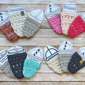 May include: Assortment of crocheted cup sleeves in various colors, including beige, brown, pink, red, teal, and black. The sleeves are designed to fit around a standard-sized cup and are made with a textured crochet stitch. The sleeves are displayed on a wooden surface with printed cards that say "To-Go Cup Sleeve" and "Cold Drink Sleeve".