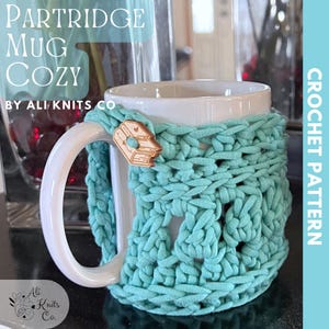 Op de afbeelding: Een turquoise blauwe gehaakte mokwarmer met een witte keramische mok. De mokwarmer heeft een hartvormig ontwerp en is bevestigd aan de mok met een houten knoop waarop "Love" staat. De tekst "PARTRIDGE MUG COZY BY ALI KNITS CO" staat in de linkerbovenhoek van de afbeelding. De tekst "CROCHET PATTERN" staat in de rechterbovenhoek van de afbeelding.