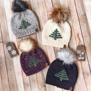 May include: Four knitted winter hats with faux fur pom-poms. Each hat features a green tree design. Colors include gray, cream, purple, and navy blue. One tag says "Let it snow" and another has snowflake designs. The hats are displayed on a wooden surface.