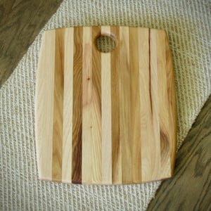 May include: A rectangular wooden cutting board constructed from vertical strips of light and medium-toned wood. The board features a smooth, polished surface with natural grain patterns and a circular cutout hole positioned near the top center edge.