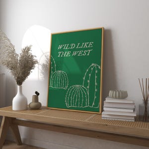 May include: A framed art print with a green background and gold frame. The print features white illustrations of cacti and the text "WILD LIKE THE WEST". The artwork is displayed on a wooden bench with decorative items.