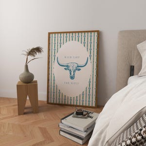 May include: A framed art print featuring a teal bull illustration with the text "WILD LIKE THE WEST" on a beige background. The print has a brown frame and is displayed in a room with a wooden side table and a vase with dried plants.