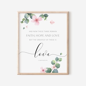 May include: A framed print with the words "AND NOW THESE THREE REMAIN FAITH, HOPE AND LOVE BUT THE GREATEST OF THESE IS love" in black script. The print is decorated with pink flowers and green leaves.