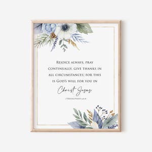 May include: A framed print with a light wood frame. The print features the text "Rejoice Always, Pray Continually, Give Thanks..." in black script. Floral watercolor accents in blue, white, and gold decorate the top and bottom corners.