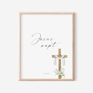 May include: A framed print featuring the words "Jesus wept" in elegant script above a wooden cross draped with white fabric. A watercolor illustration of white lilies and greenery is at the base of the cross. The text "JOHN 11:35" is at the bottom.