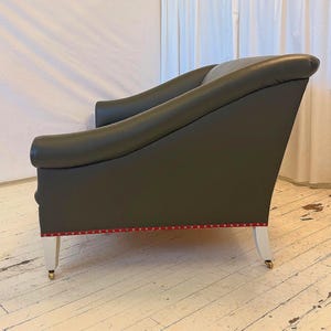 Puede incluir: Un sillón gris oscuro con brazos curvos y respaldo alto. La silla tiene patas blancas con ruedas doradas. Un ribete rojo con pequeños tachuelas doradas acentúa la base de la silla.