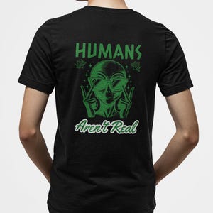 Op de afbeelding: Zwart t-shirt met een groene afbeelding van een alien met een zonnebril en een vredesteken. De tekst "HUMANS Aren't Real" staat boven en onder de illustratie.