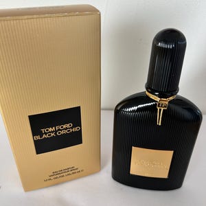 May include: A black Tom Ford Black Orchid perfume bottle and matching box. The bottle has a ribbed design and a gold accent. The box is gold with the brand and product name in black.