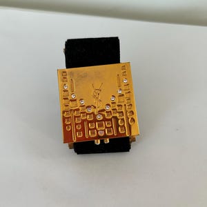 Könnte beinhalten: Goldfarbene rechteckige Brosche mit dekorativem Muster und eingebetteten Strasssteinen. Die Brosche hat ein Logo in der Mitte und ist auf einem schwarzen Samtrücken befestigt.