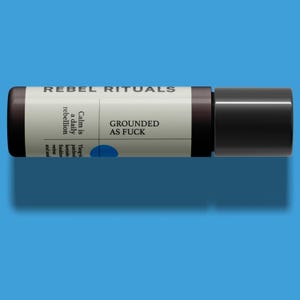 May include: A cylindrical product with a black cap and a label that reads "REBEL RITUALS" and "GROUNDED AS FUCK." The label also includes text about calm and rebellion, with a blue circle accent. The product is set against a blue background.