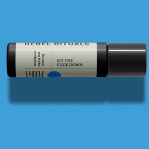 May include: A black and white cylindrical product with the text "REBEL RITUALS" at the top. The product has the text "Because rest is the revolution" and "SIT THE FUCK DOWN". A blue dot is visible on the product.
