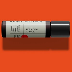 May include: A cylindrical product with a black cap and a label that reads "REBEL RITUALS" and "HORMONAL AS FUCK". The label also includes text about the product's use and a red circle. The product is set against an orange background.