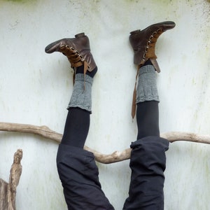 May include: A pair of brown leather boots with grey knitted leg warmers. The boots are worn by a person standing on their hands with their legs in the air. The person is leaning against a white wall with a piece of driftwood behind them.