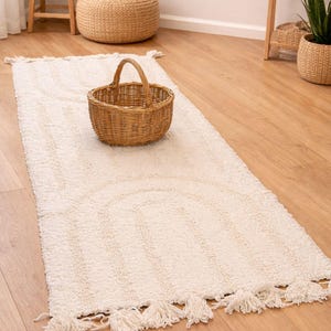 May include: A cream-coloured, rectangular rug with a tufted design and tassels at the ends. A woven basket with a handle sits on the rug. The rug has a subtle arched pattern. The scene is set on a wooden floor.