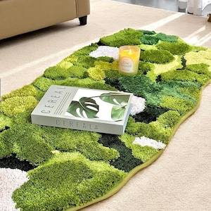 Pode incluir: Um tapete verde em forma de musgo com diferentes tons de verde e manchas brancas. Um livro intitulado "CEREAL" com um design de folha de monstera e uma vela acesa estão sobre o tapete. O tapete tem uma forma arredondada e orgânica.