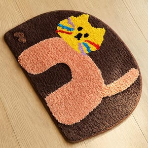 Peut inclure: Un tapis marron semi-circulaire avec un motif de chat de dessin animé. La tête du chat est jaune avec des accents colorés, et le corps est pêche. Le tapis est fait d'un matériau doux et texturé, idéal pour une chambre d'enfant ou une salle de bain.