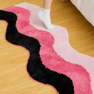 May include: A wavy rug in shades of pink and black. The rug has a soft, textured surface and a unique, flowing design. The rug is placed on a wooden floor.