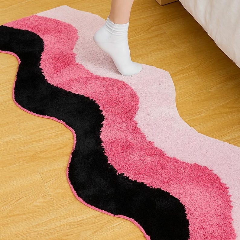 May include: A wavy rug in shades of pink and black. The rug has a soft, textured surface and a unique, flowing design. The rug is placed on a wooden floor.