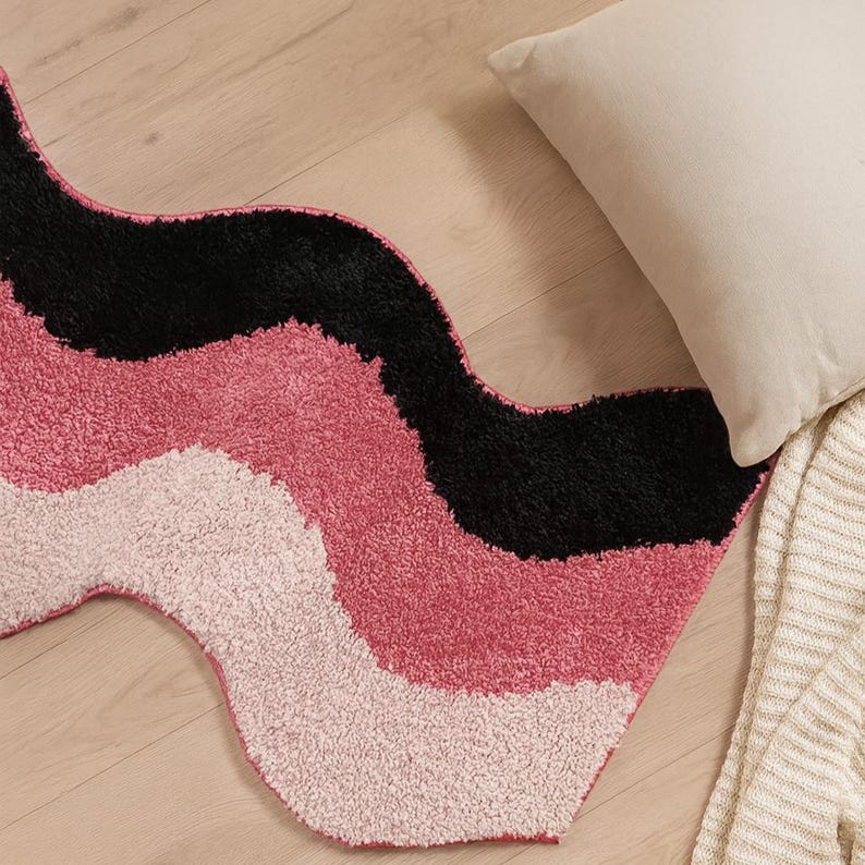 May include: A wavy rug with a textured surface, featuring bands of black, pink, and light pink. A beige pillow and a cream-colored blanket are visible in the background. The rug is placed on a light wood floor.