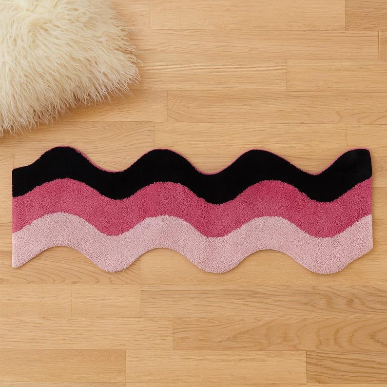 May include: A wavy, rectangular rug with a black, medium pink, and light pink color scheme. The rug is made of a soft, plush material and is placed on a wooden floor. A fluffy, white pillow is in the background.