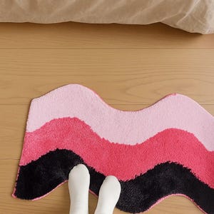 May include: A wavy, multi-colored rug with a soft, textured surface. The rug features bands of pink, dark pink, and black. The rug is placed on a wooden floor, with a beige sheet visible in the background. The rug is a decorative home accent.
