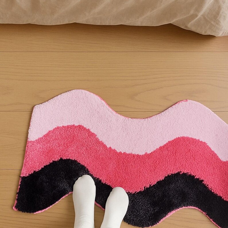 May include: A wavy, multi-colored rug with a soft, textured surface. The rug features bands of pink, dark pink, and black. The rug is placed on a wooden floor, with a beige sheet visible in the background. The rug is a decorative home accent.