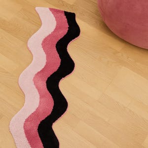 May include: A wavy rug with a color block design. The rug features stripes of light pink, medium pink, and black. The rug is on a light wood floor. A pink, round, velvet ottoman is in the background.