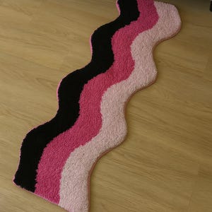 May include: A wavy rug with a black, pink, and light pink striped design. The rug has a soft, textured surface and a pink border. The rug is placed on a wooden floor.