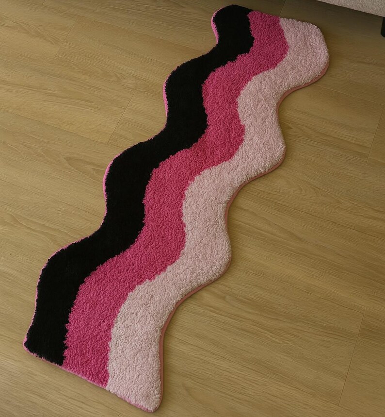 May include: A wavy rug with a black, pink, and light pink striped design. The rug has a soft, textured surface and a pink border. The rug is placed on a wooden floor.